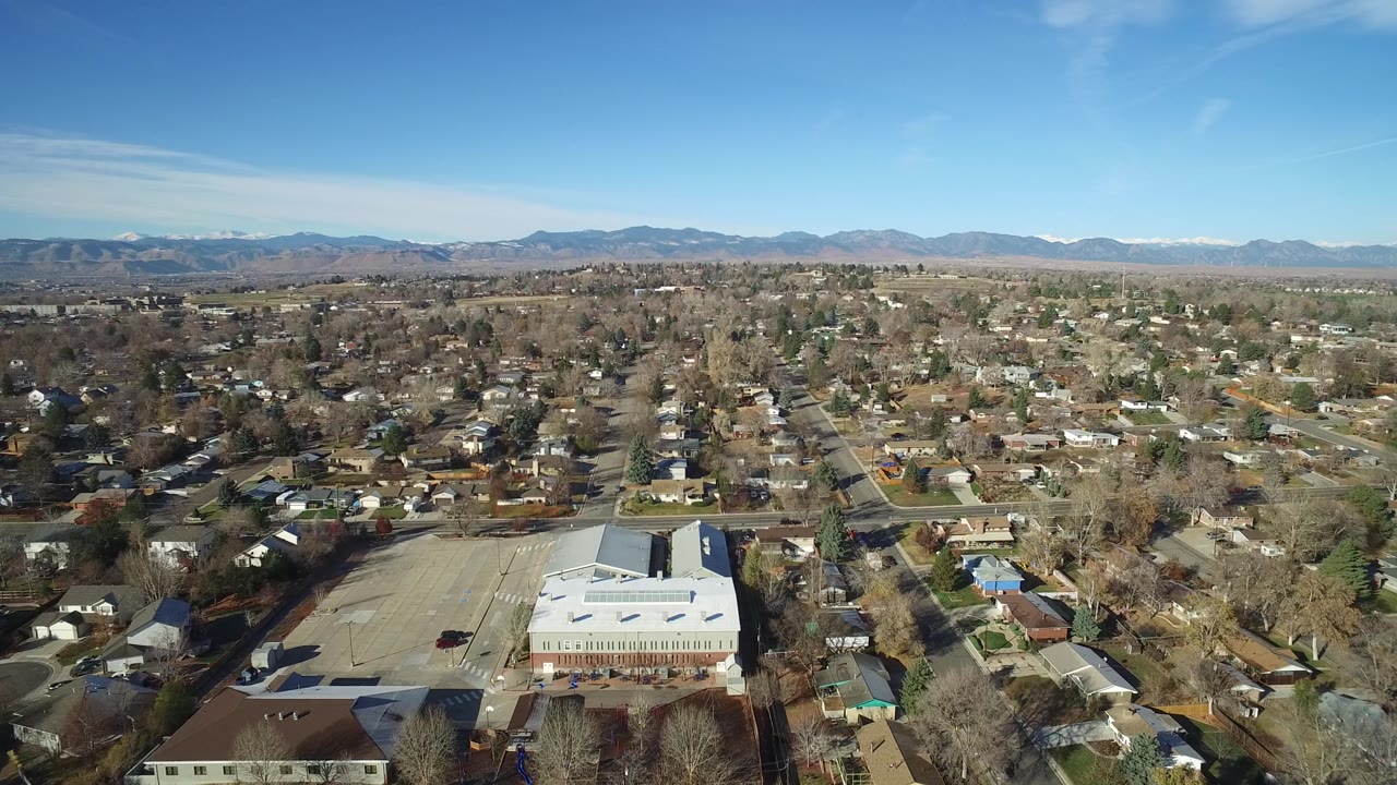 Second frame of the Westminster aerial holds on the neighborhood grid, with the large light-roofed building lower center, houses and bare trees across the midground, and no visible people.