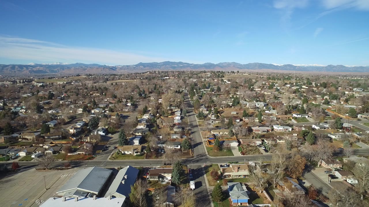 Wide aerial view of a Westminster, Colorado neighborhood with houses, bare trees, gridded streets, blue sky, and distant mountains, with no visible people.