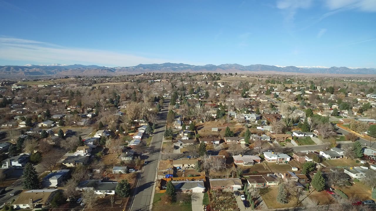 Wide aerial view over Westminster, Colorado residential blocks with houses, leafless trees, evergreen trees, distant mountains, and no visible people.