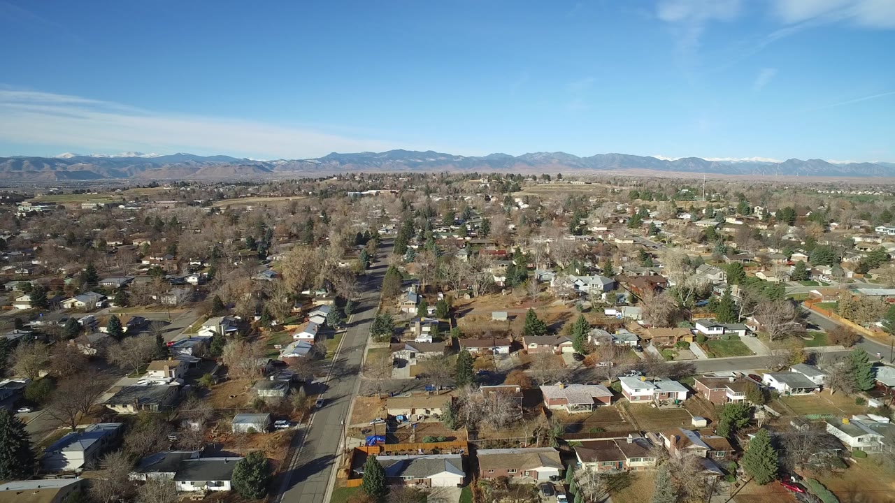 Drone-like wide view of houses and tree-lined roads with mountains beyond the city.