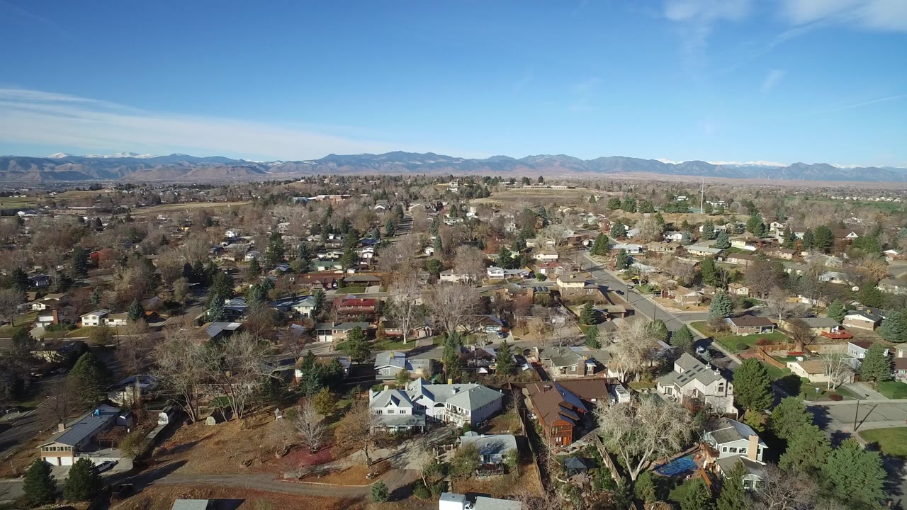Early frame of Westminster, Colorado from above, showing detached homes, tree-lined streets, bare branches, lawns, and distant mountains under a clear blue sky with no visible people.