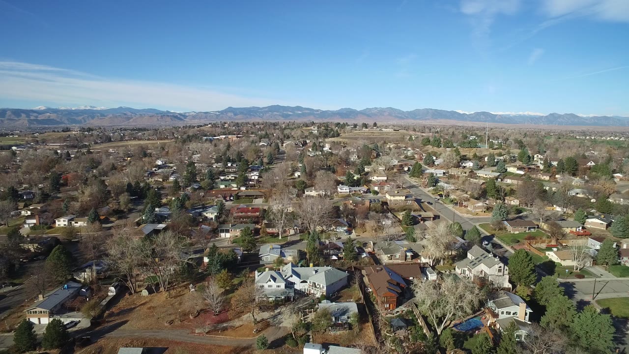 Aerial frame over Westminster, Colorado with houses and yards filling the lower frame, a straight residential road near center, open land beyond the neighborhood, and no visible people.
