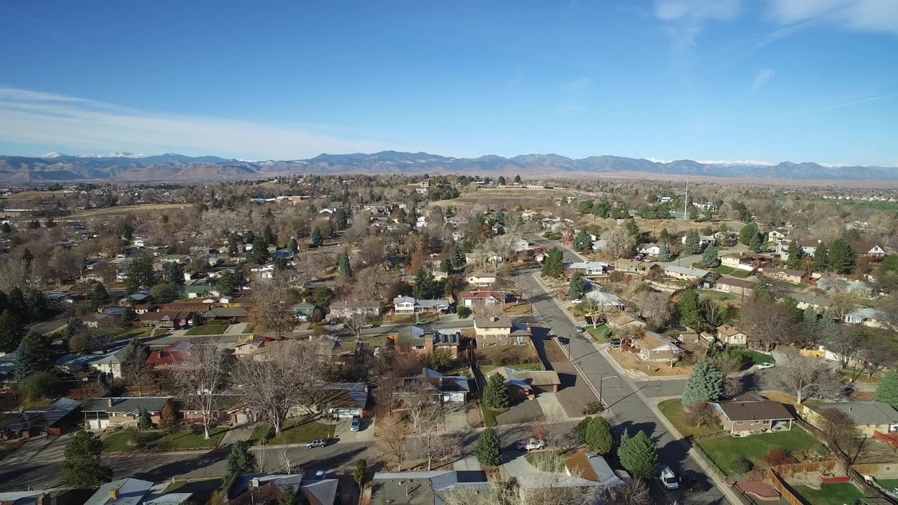 Westminster, Colorado neighborhood seen from above with rooftops, a central street corridor, winter-bare trees, and distant mountains under clear daylight, with no visible people.