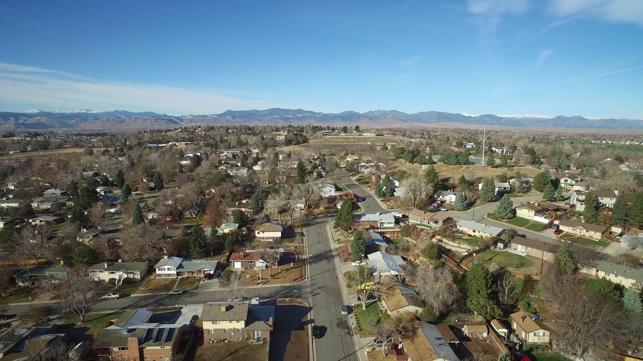 Aerial view over Westminster, Colorado suburban homes, neighborhood streets, bare trees, green lawns, and distant mountains under a blue sky, with no visible people.