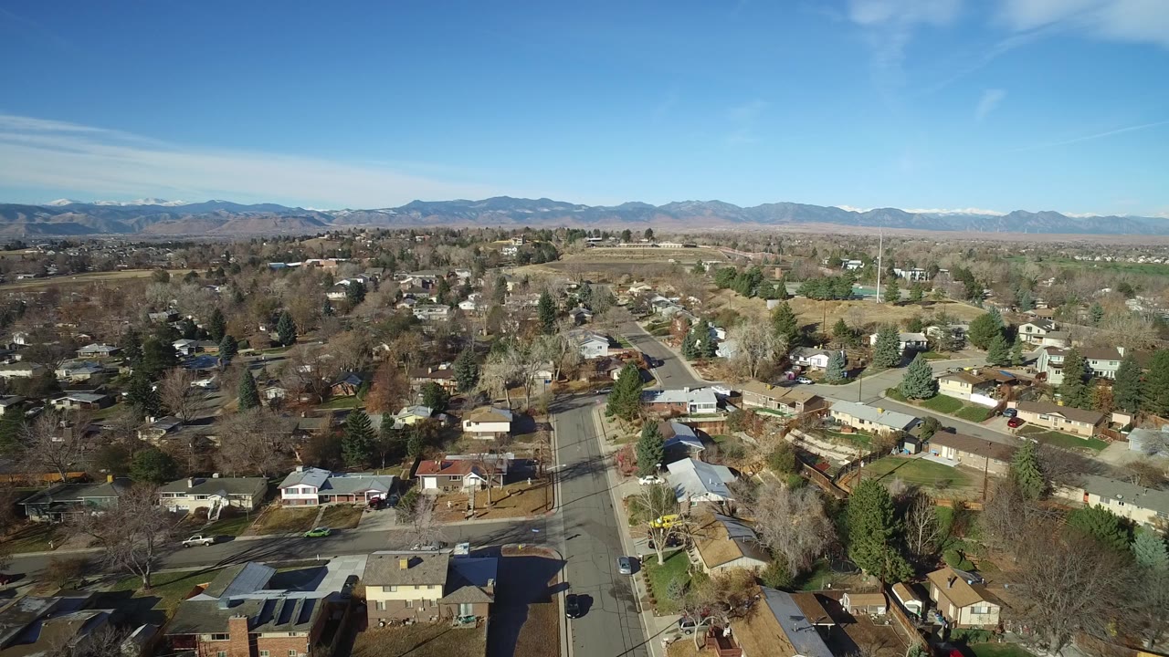 High aerial frame of Westminster, Colorado showing a central residential street, rows of houses, mixed green and brown yards, bare trees, and mountains on the horizon, with no visible people.