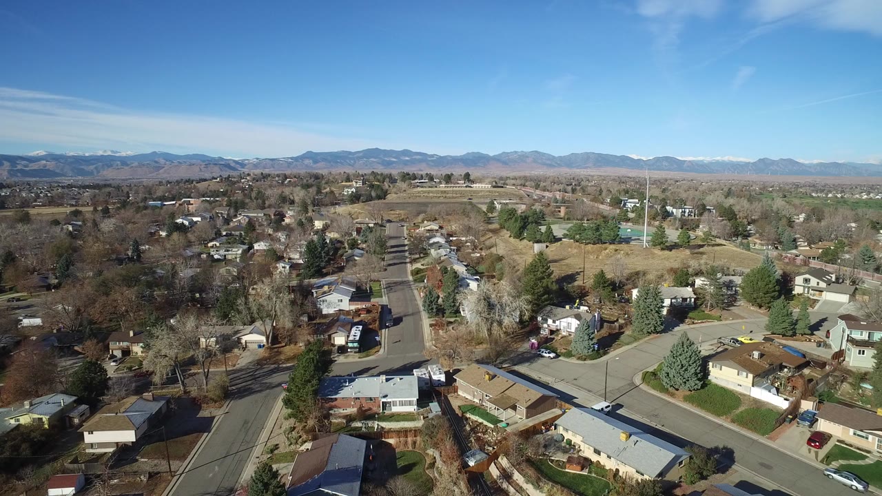 Aerial view over suburban streets, houses, trees, open land, and distant mountains.