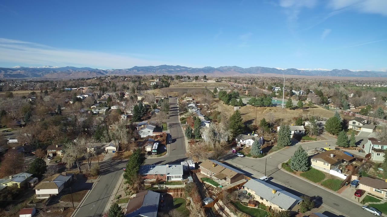 High aerial view across Westminster, Colorado homes and neighborhood roads, with a broad dry field and a mountain range on the horizon, and no visible people.