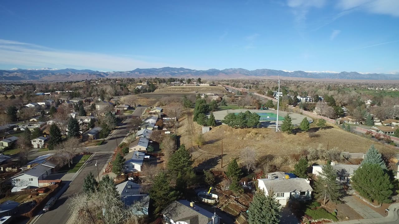 Elevated Westminster, Colorado view with rooftops in the lower frame, a road and tennis courts near center, winter trees across the neighborhood, and distant mountains under clear sky.