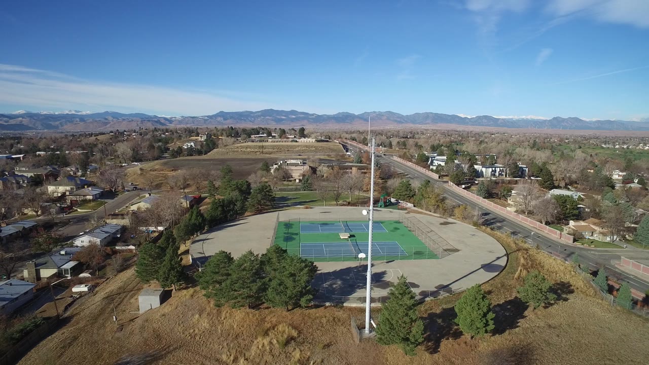 Aerial frame in Westminster, Colorado showing the tennis courts slightly farther across the frame, with a road leading toward neighborhoods, open land, mountains, and no visible people.