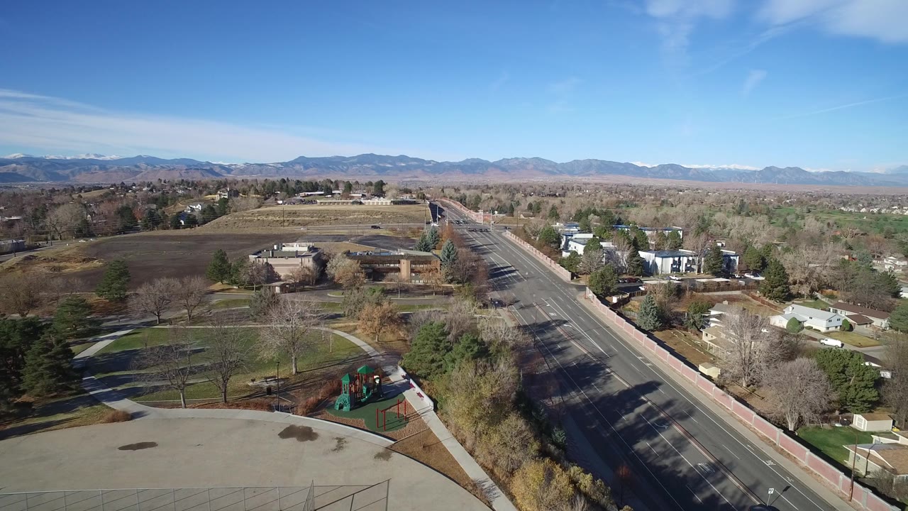 Westminster, Colorado aerial frame looking farther along the road, with homes and small buildings on both sides, open fields beyond, distant mountains, and no visible people.