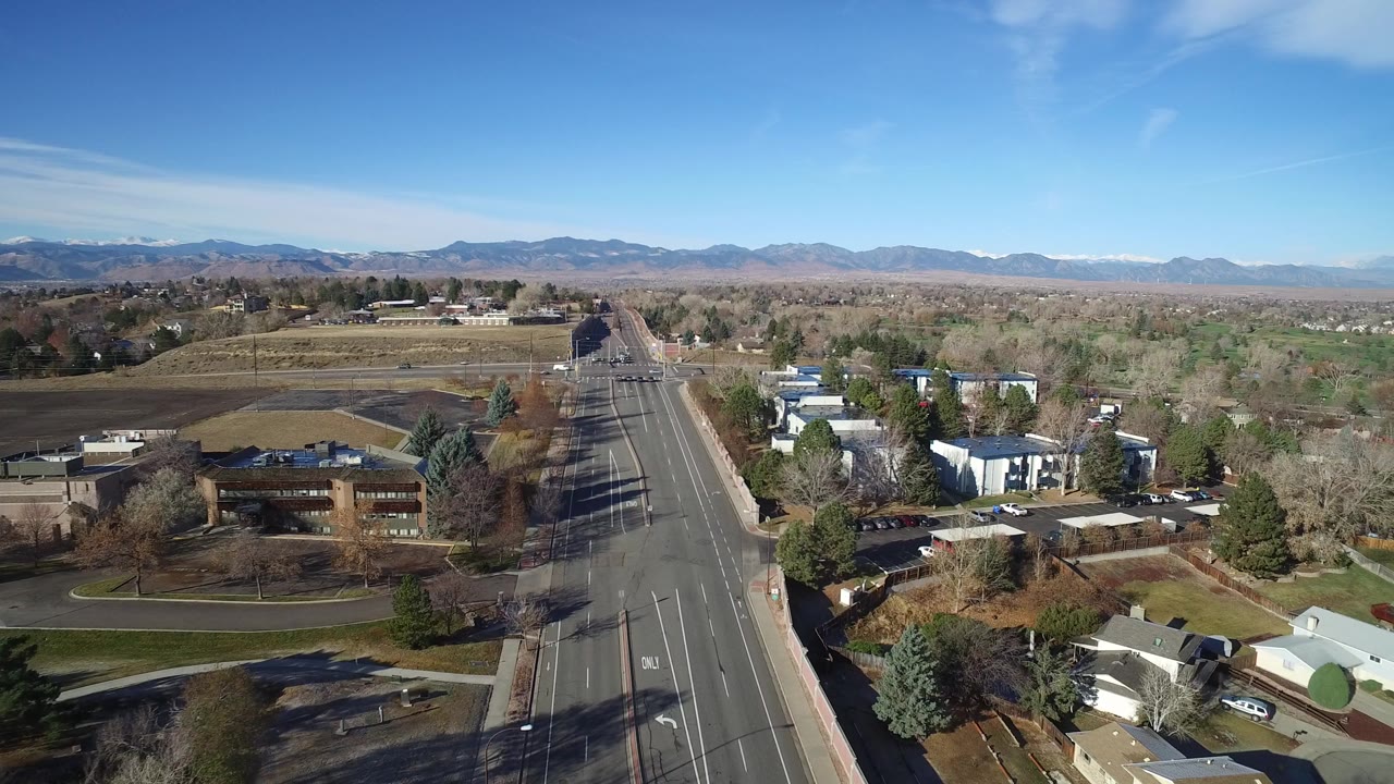 Early frame of Westminster, Colorado from above, showing a straight multi-lane road, scattered buildings, leafless trees, distant mountains, clear blue sky, and no visible people.