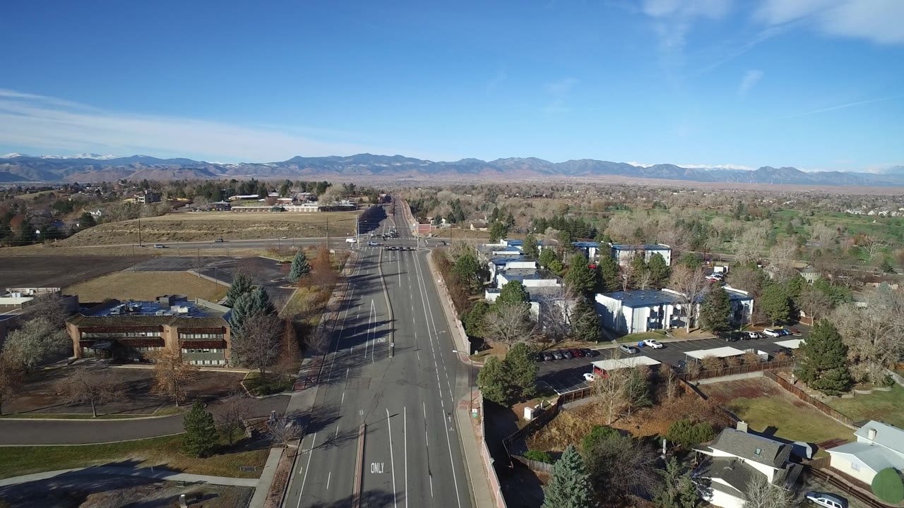 Aerial city edge view with road, parking lots, trees, and mountains.