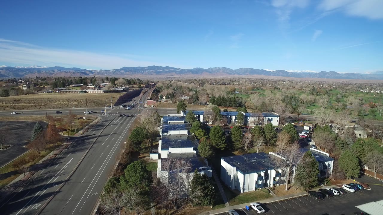 Aerial view in Westminster, Colorado showing the building cluster lower in frame, the main road at left, scattered parked cars, green and leafless trees, distant mountains, and no visible people.