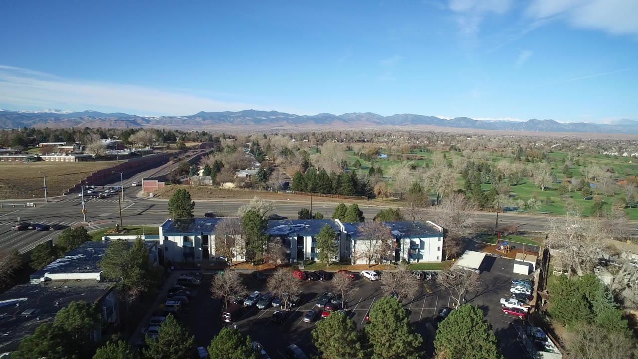 High aerial view in Westminster, Colorado showing low buildings and parked cars in the foreground, a road and green tree cover beyond, distant mountains, and no visible people.