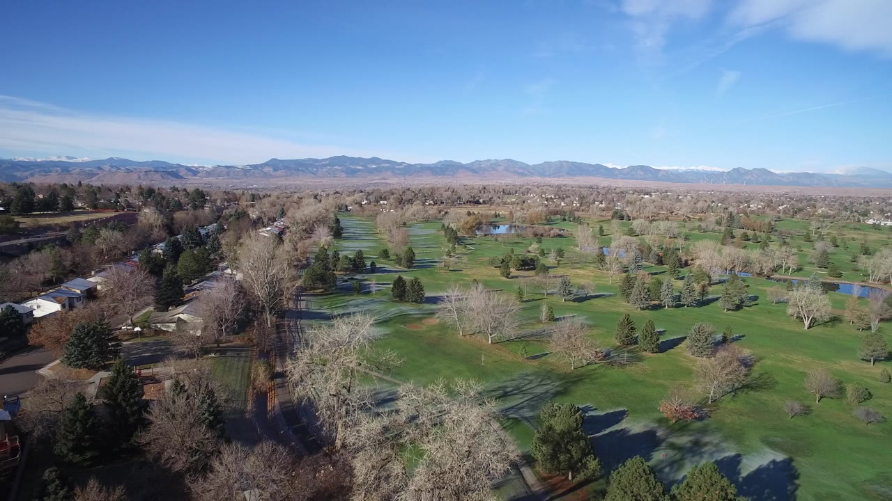 Aerial view over green fields, trees, ponds, houses, and distant mountains.