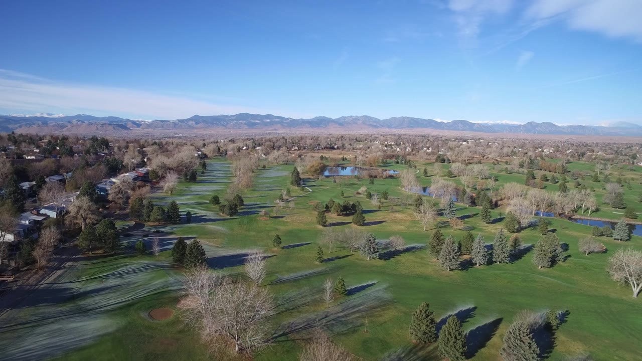 Aerial view in Westminster, Colorado with the green course-like landscape filling the foreground, ponds and tree lines through the center, houses along the left edge, mountains on the horizon, and no visible people.