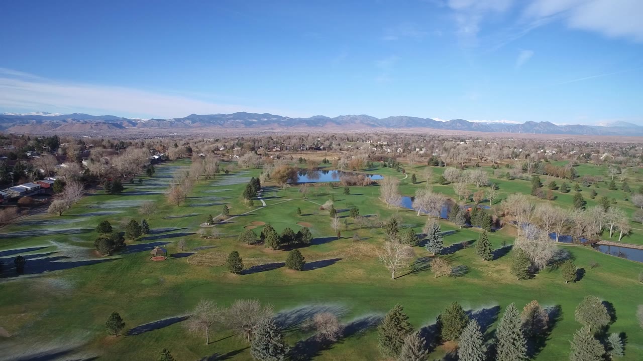 Wide aerial view in Westminster, Colorado showing green fields, scattered trees, small water areas, distant neighborhoods, mountains, blue sky, and no visible people.