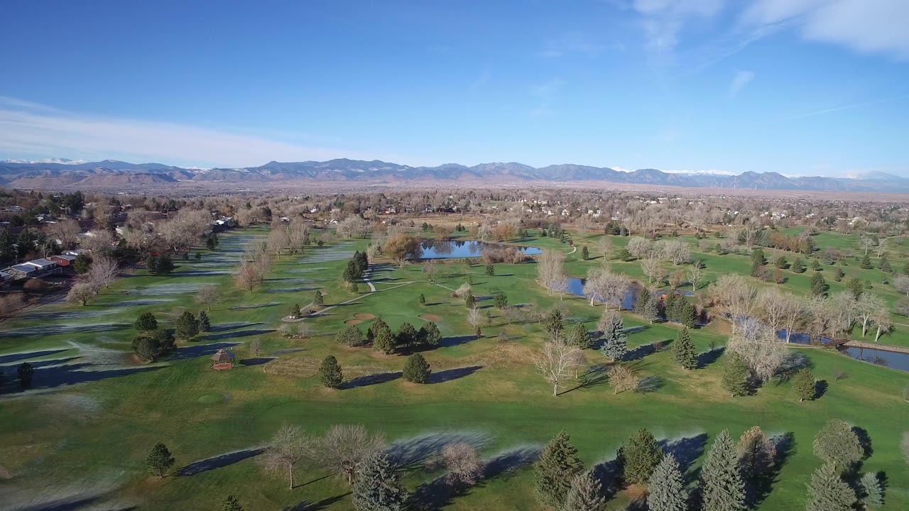 Aerial frame over Westminster, Colorado with fairway-like grass, leafless trees, reflective ponds, distant homes, mountains, clear sky, and no visible people.