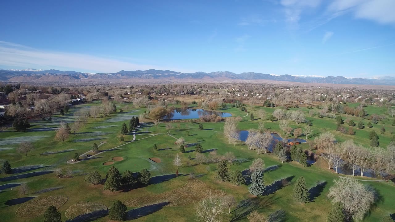 A wide aerial view in Westminster, Colorado shows green fairways, scattered trees, ponds, and a distant mountain horizon under a blue sky, with no visible people.