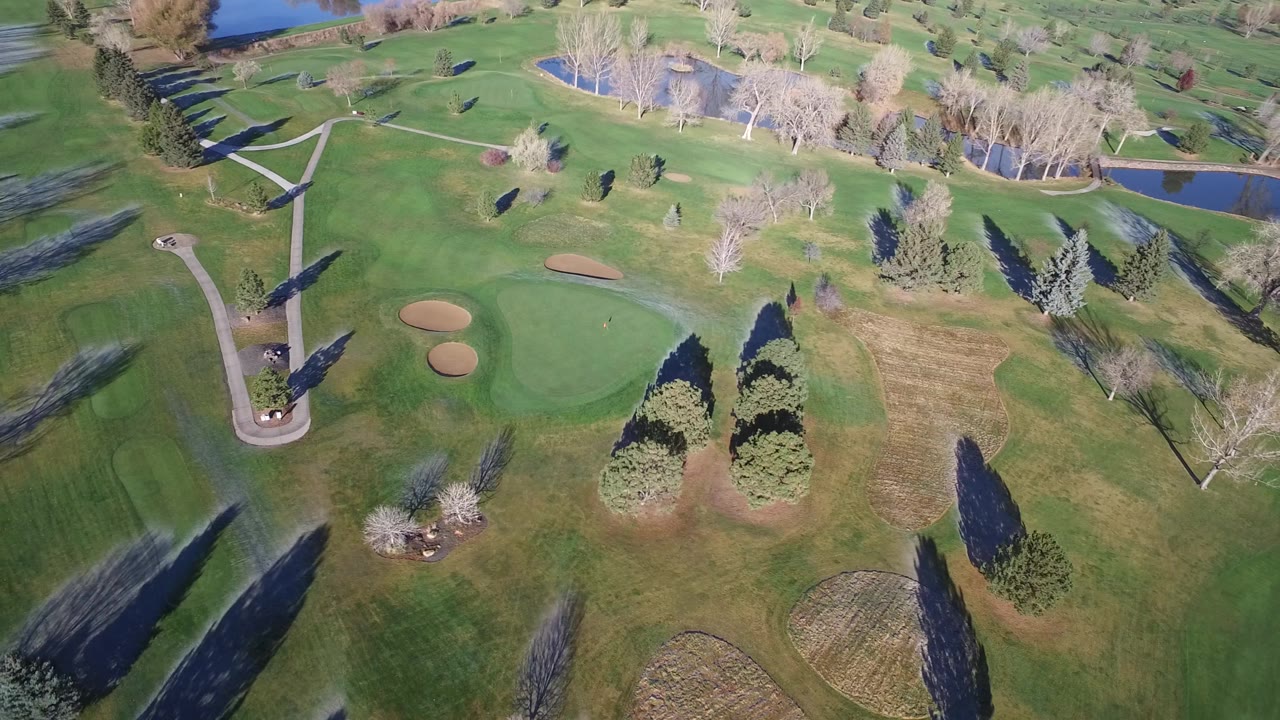 Aerial frame over Westminster, Colorado showing a paved path bending through open grass, two tan bunkers near the green, ponds in the background, and no visible people.