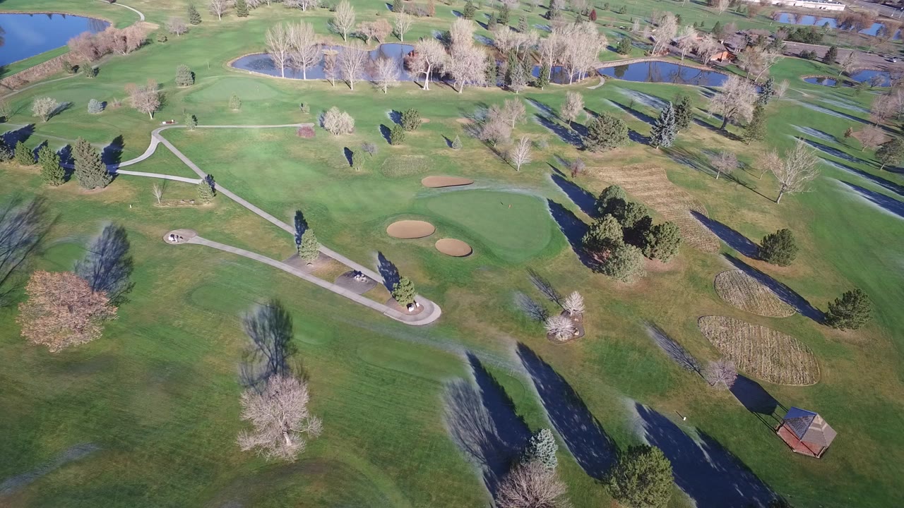 A high aerial frame over Westminster, Colorado green grounds shows curved paved paths, tan sand bunkers, ponds near the top edge, scattered trees, long shadows, and no visible people.