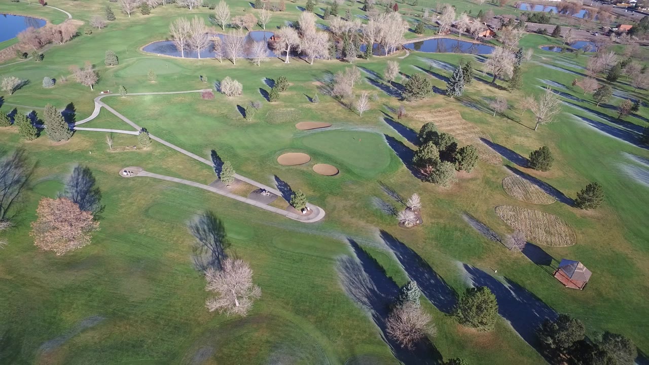 Aerial landscape with fairway-like grass, trees, paths, bunkers, and water.