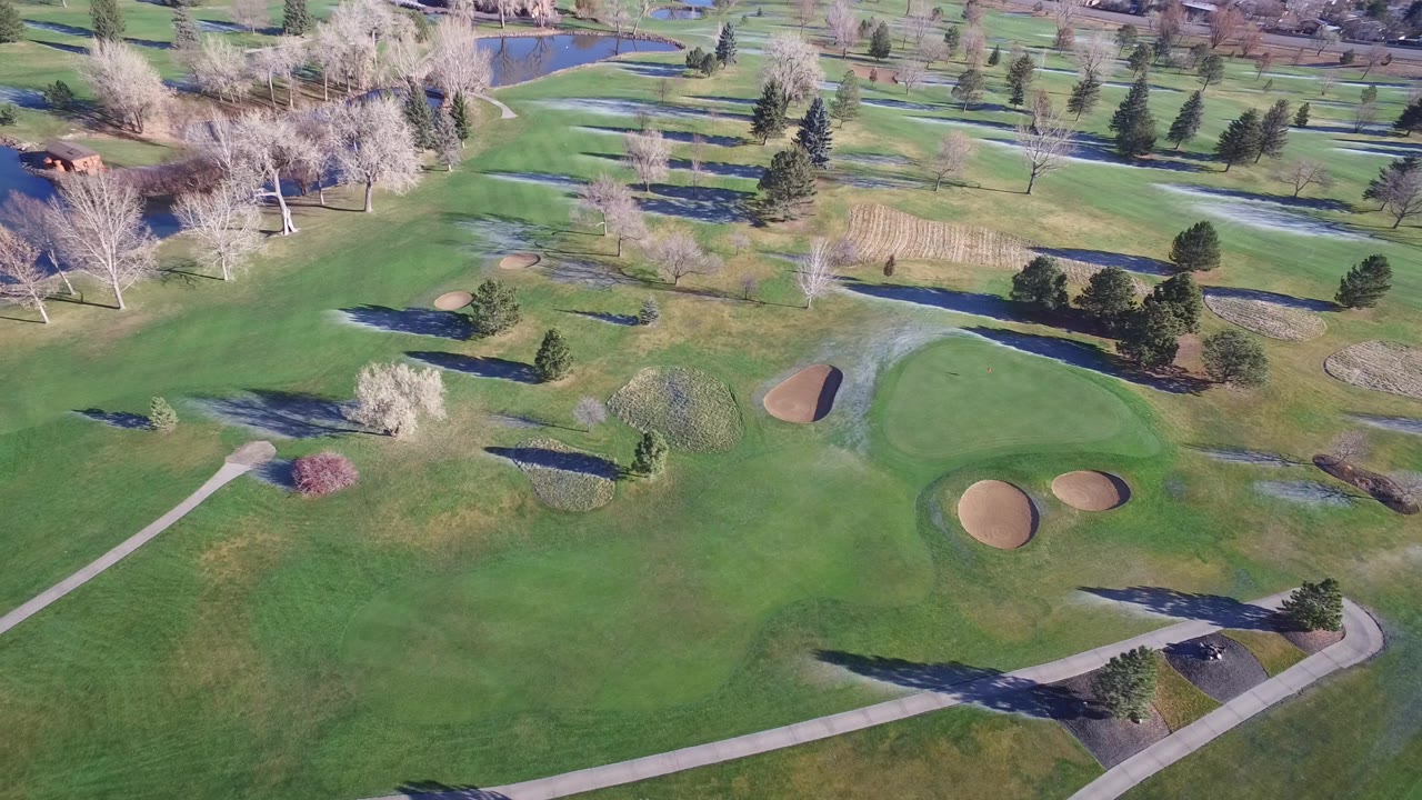 Aerial frame in Westminster, Colorado showing a broad fairway, paired sand bunkers, pale cart paths, scattered trees, reflective water patches, and no visible people.