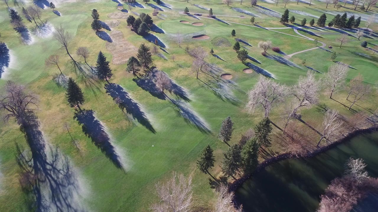 Overhead frame from Westminster, Colorado showing green fairways, scattered trees, sand bunkers, paved paths, and a dark pond edge with no visible people.