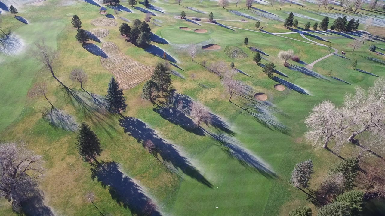 Overhead view in Westminster, Colorado showing green fairway-like turf, sand bunkers, scattered trees, long shadows, and small snow patches, with no visible people.