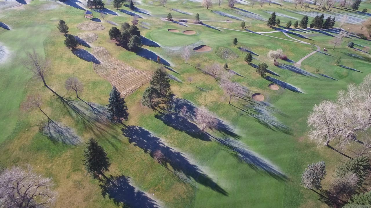 Aerial view of landscaped greens with bunkers, trees, and water at the edge.
