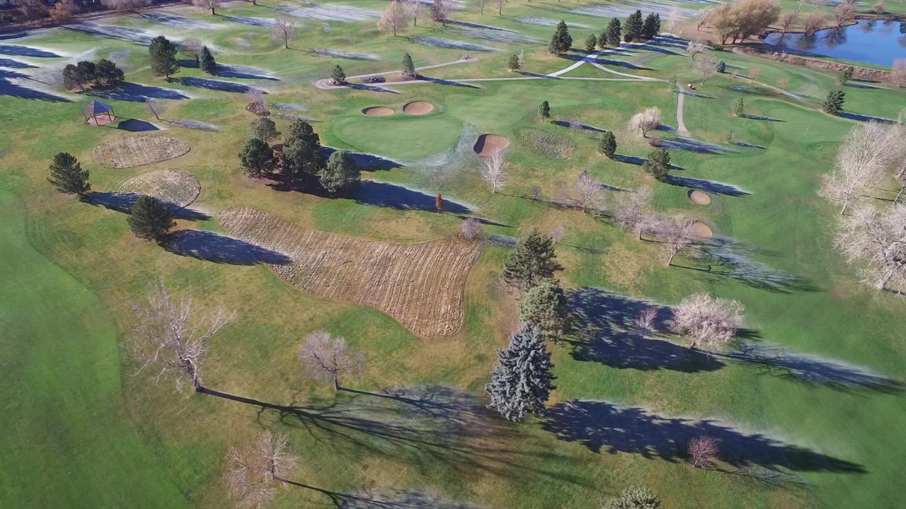 A slightly shifted aerial view of the Westminster, Colorado grounds shows clustered evergreens, bare deciduous trees, tan patches, winding paths, distant ponds, and no visible people.