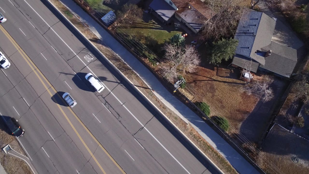 Overhead aerial frame in Westminster, Colorado showing a multi-lane road, sidewalks, rooftops, yards, bare trees, cars, and no clearly visible people at the start of the clip.