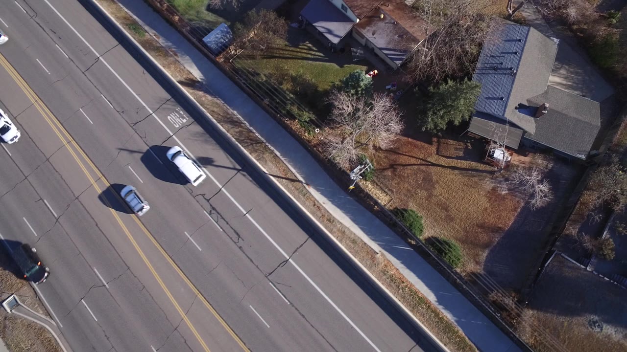 Aerial frame over Westminster, Colorado with the road still dominant on the left, a white car on the pavement, detached homes to the right, fenced yards, trees, and no clearly visible people.