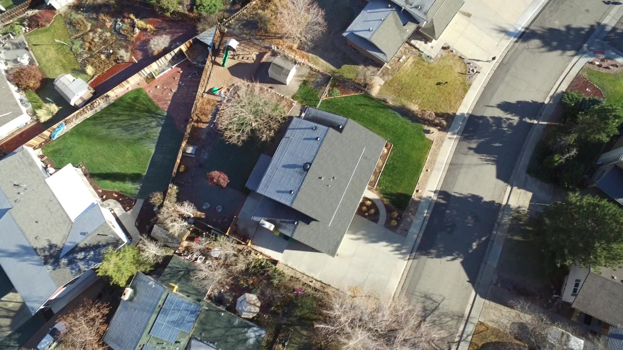 Overhead aerial frame in Westminster, Colorado showing gray-roofed homes, fenced yards, green lawns, driveways, and a quiet street with no visible people.