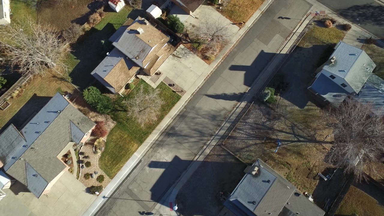 Aerial frame over Westminster, Colorado homes, showing gray and tan rooftops, driveways, winter-bare trees, lawns, parked vehicles, and no clearly visible people.