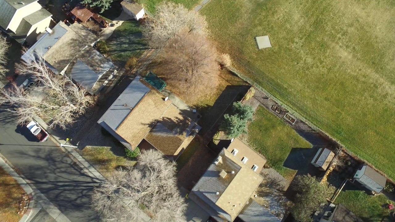 Aerial frame over Westminster, Colorado homes beside a large green field, with roofs, driveways, leafless trees, parked cars, and no visible people.