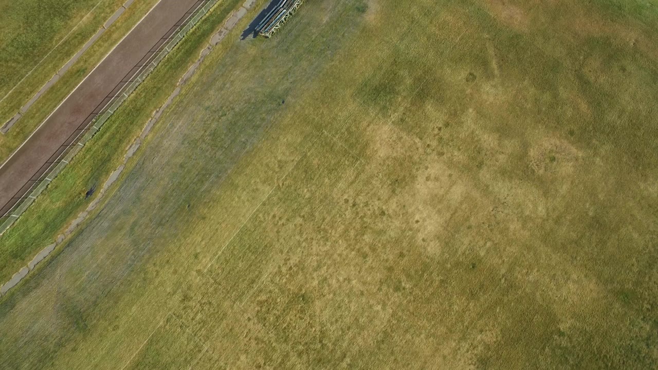 Early overhead frame in Westminster, Colorado showing mostly grass, a diagonal paved edge, and small metal bleachers near the top of frame, with no visible people.