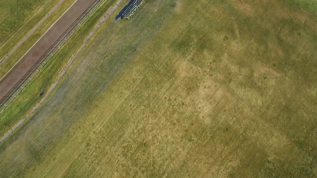 Overhead Westminster, Colorado view with the grass field centered, a diagonal paved strip along the left side, and bleachers near the top edge, with no visible people.
