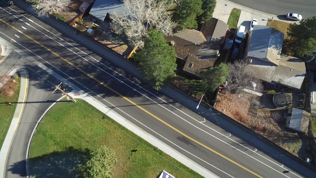 Aerial view in Westminster, Colorado with a broad asphalt road, painted lane markings, a corner lawn, rooftops, driveways, and mixed evergreen and leafless trees, with no visible people.