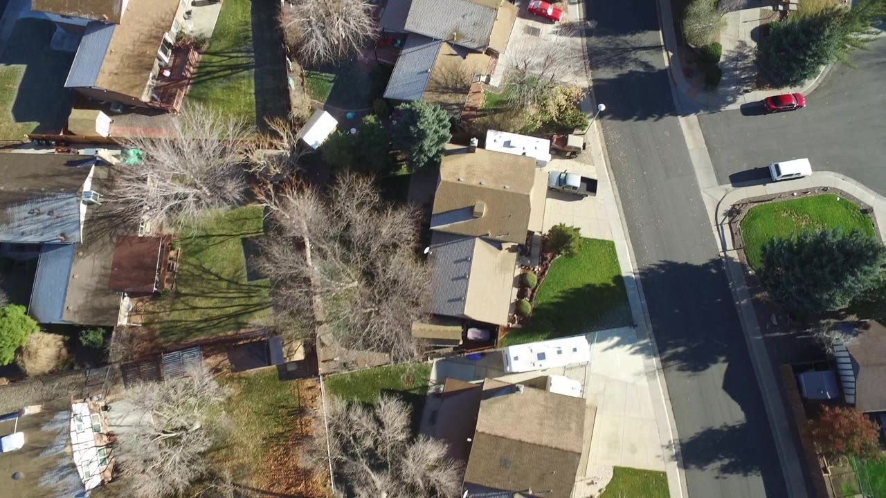 Top-down Westminster, Colorado neighborhood frame showing tan rooftops, green lawns, parked vehicles, bare trees, and sharp shadows crossing residential streets, with no visible people discernible.
