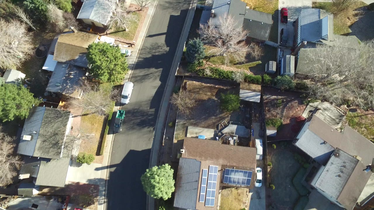 High aerial frame over Westminster, Colorado homes showing a straight residential street, rooflines, fenced yards, parked cars, strong tree shadows, and no visible people.