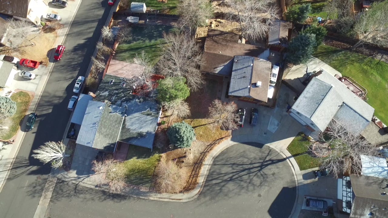Overhead view of suburban houses, yards, trees, streets, and parked cars.