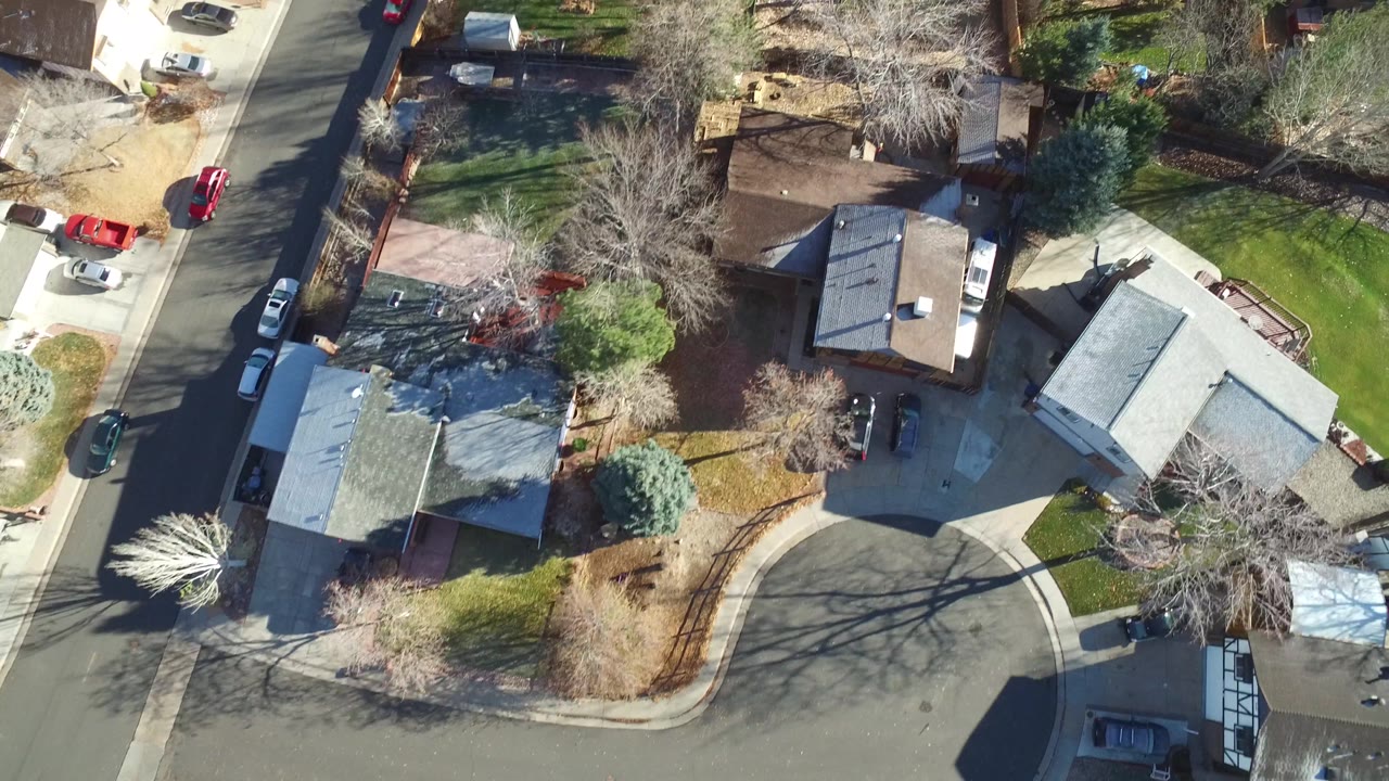 Aerial frame over Westminster, Colorado homes near a curved residential street, showing lawns, fences, driveways, several parked cars, and bare tree shadows, with no visible people.