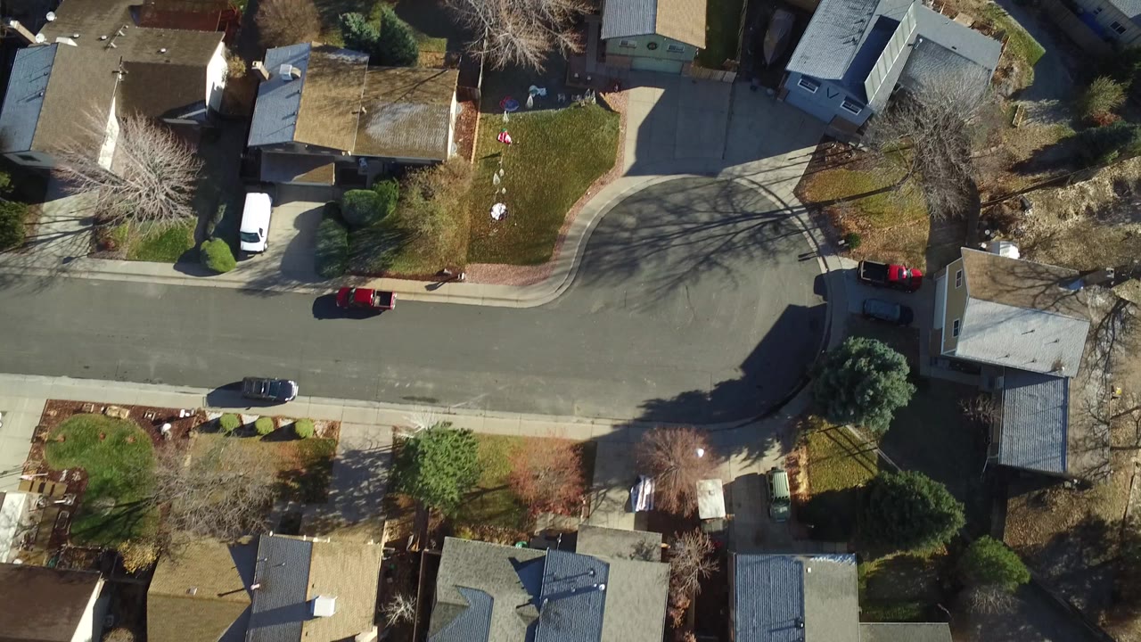 Aerial frame over Westminster, Colorado with rooftops, a broad residential road, front lawns, driveways, parked vehicles, bare trees, and no clearly visible people.