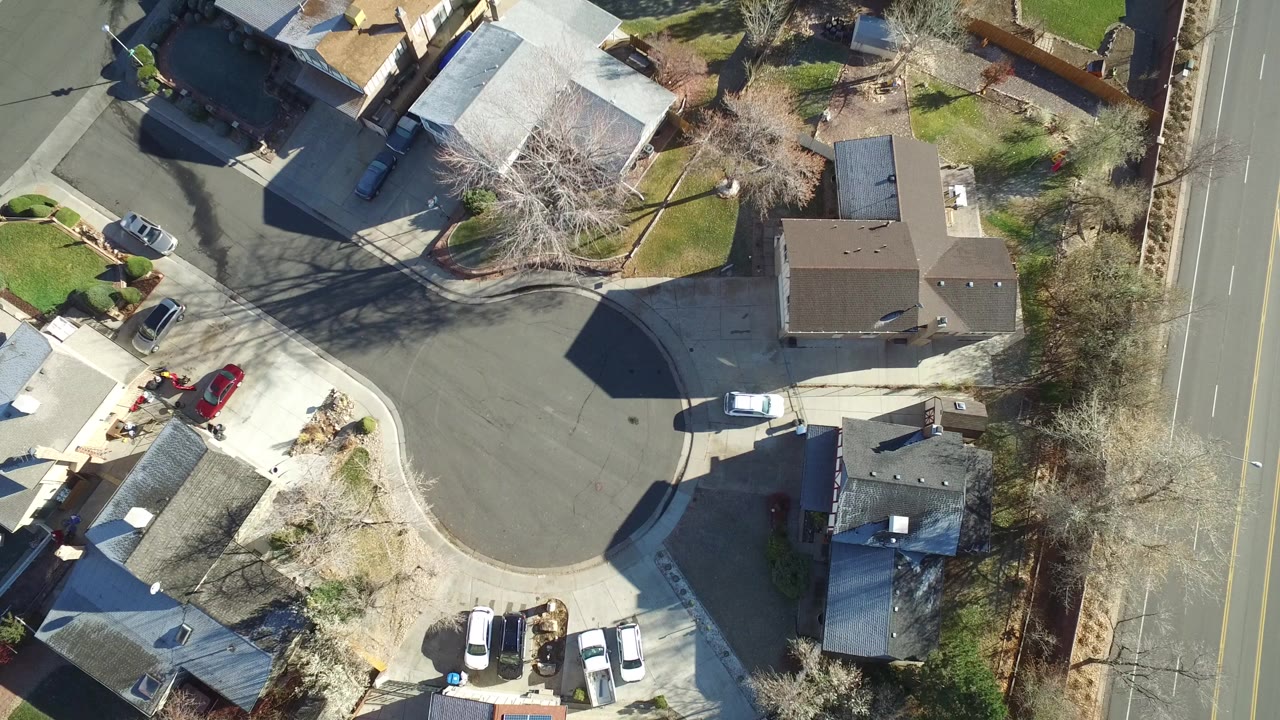 Overhead aerial frame in Westminster, Colorado showing a cul-de-sac, private homes, driveways, parked vehicles, leafless trees, and a larger road at the right edge, with no visible people.