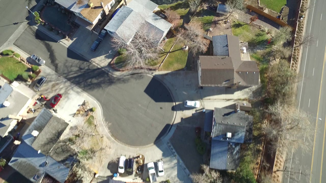 A second overhead moment in Westminster, Colorado holds on the cul-de-sac, showing rooftops, parked cars, tree shadows, small lawns, and the bordering road, with no visible people.