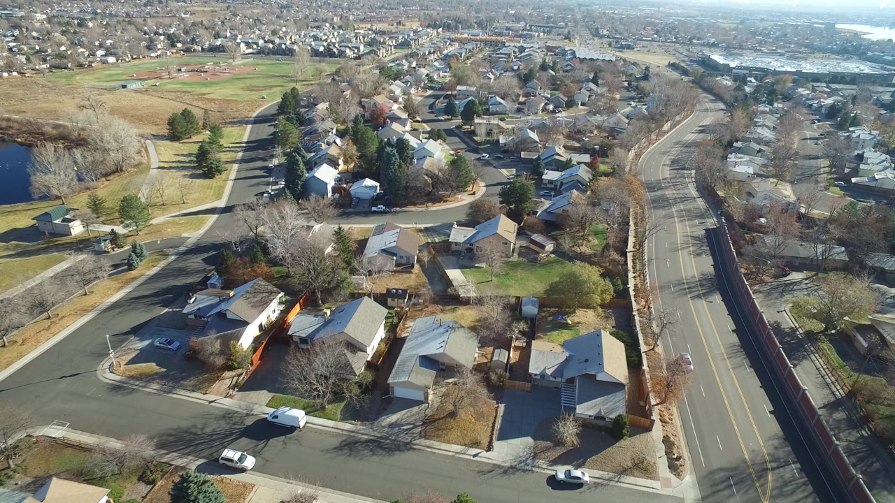High aerial frame of Westminster, Colorado residential blocks with rooftops, a curved side street, cars on neighborhood roads, open land at left, distant water, and no visible people.