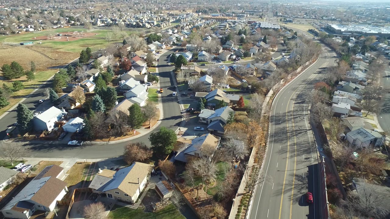 Slightly shifted aerial view of Westminster, Colorado rooftops, neighborhood streets, bare trees, evergreen trees, and a broad road, with no visible people.