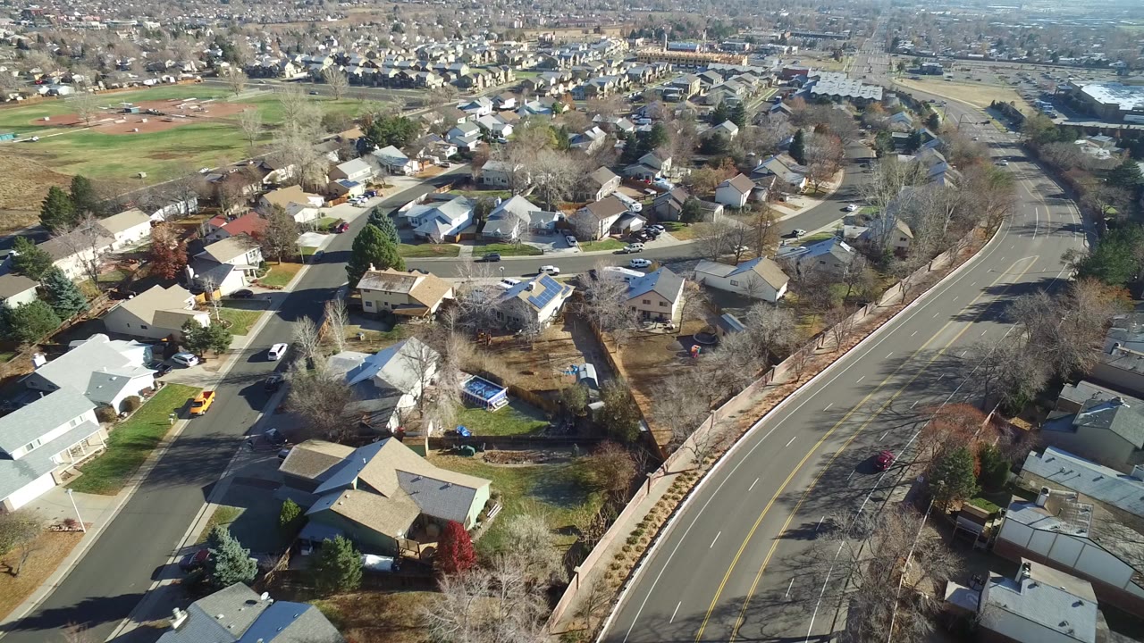 High aerial frame over Westminster, Colorado homes, yards, bare trees, and a broad curving road at the start of the clip, with no visible people.