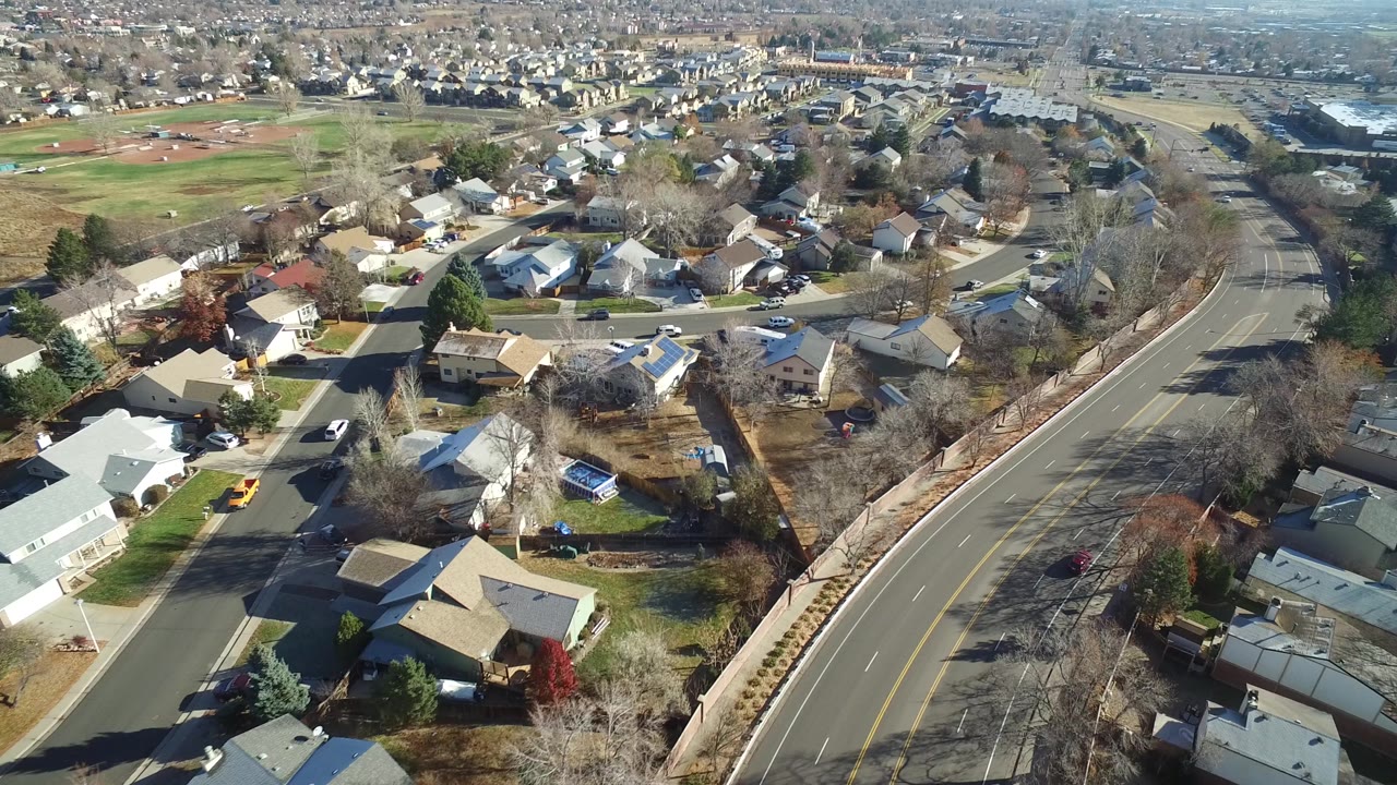 Aerial view across Westminster, Colorado residential blocks, with rooftops and yards in the foreground and the curving road running along the right side, with no visible people.